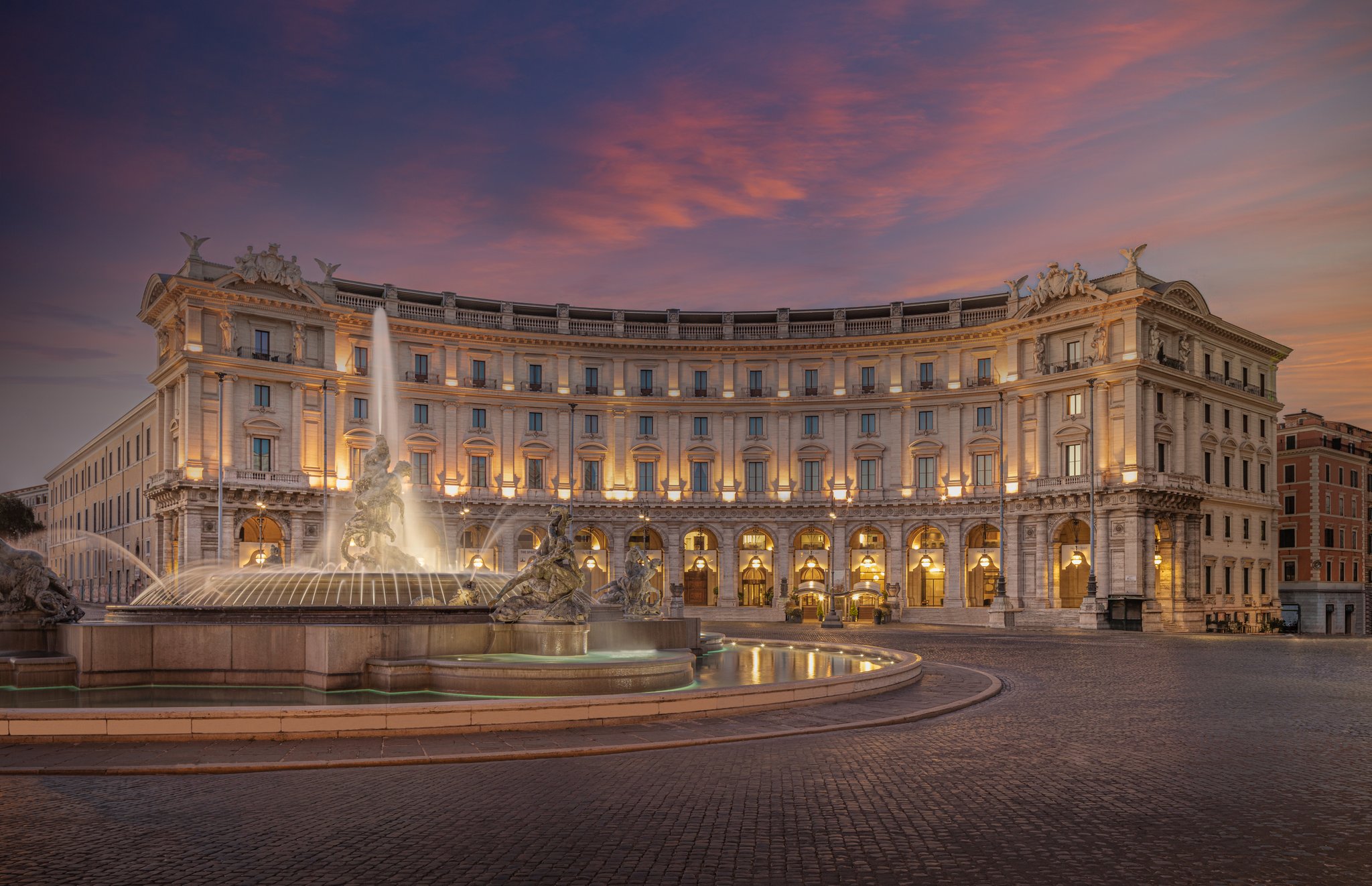 Sunset photo of a rounded building in Europe with a brick common area and a water fountain in the middle.