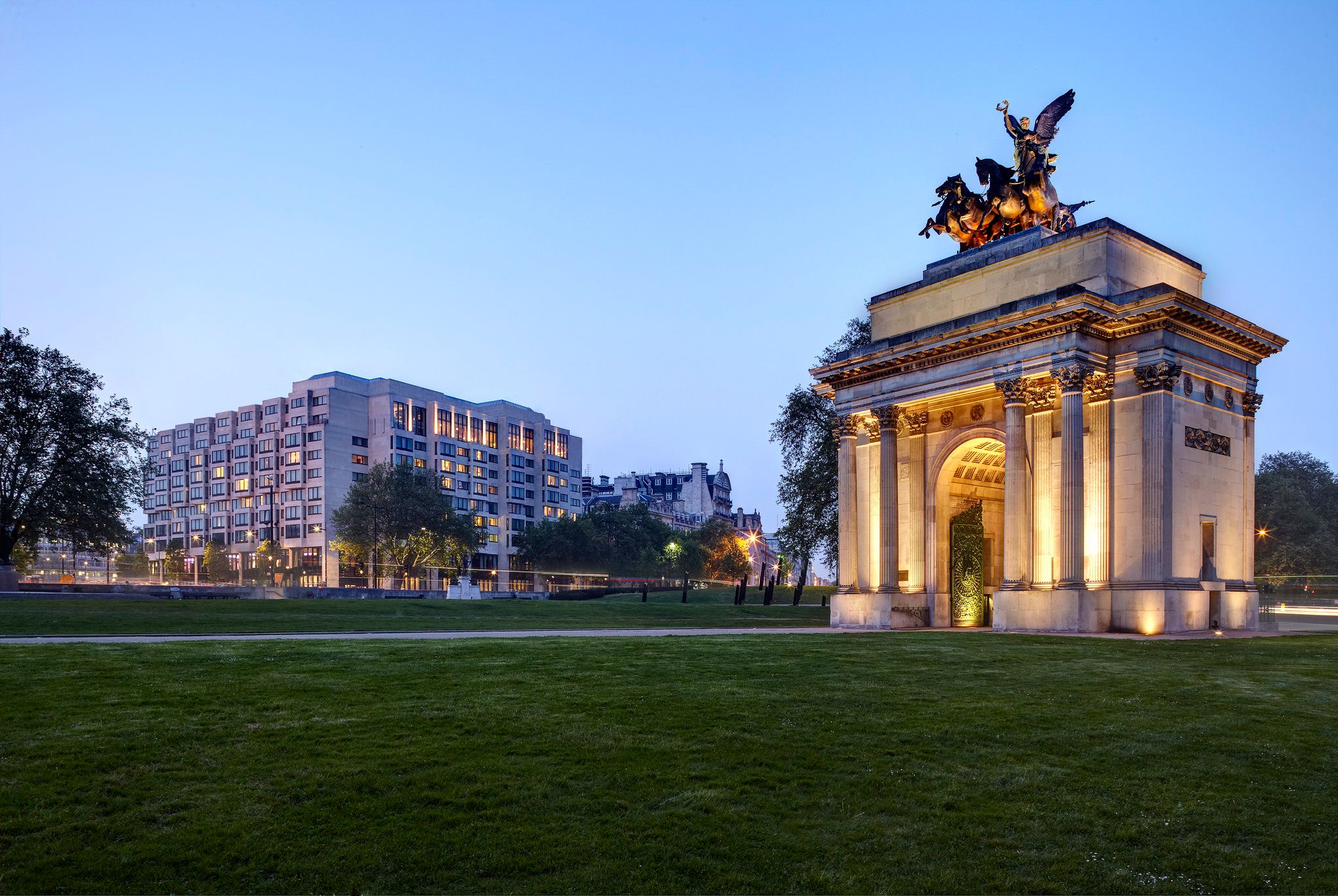 Evening picrue of hotel in background, a grassy common area and a large lit up archway in Europe.